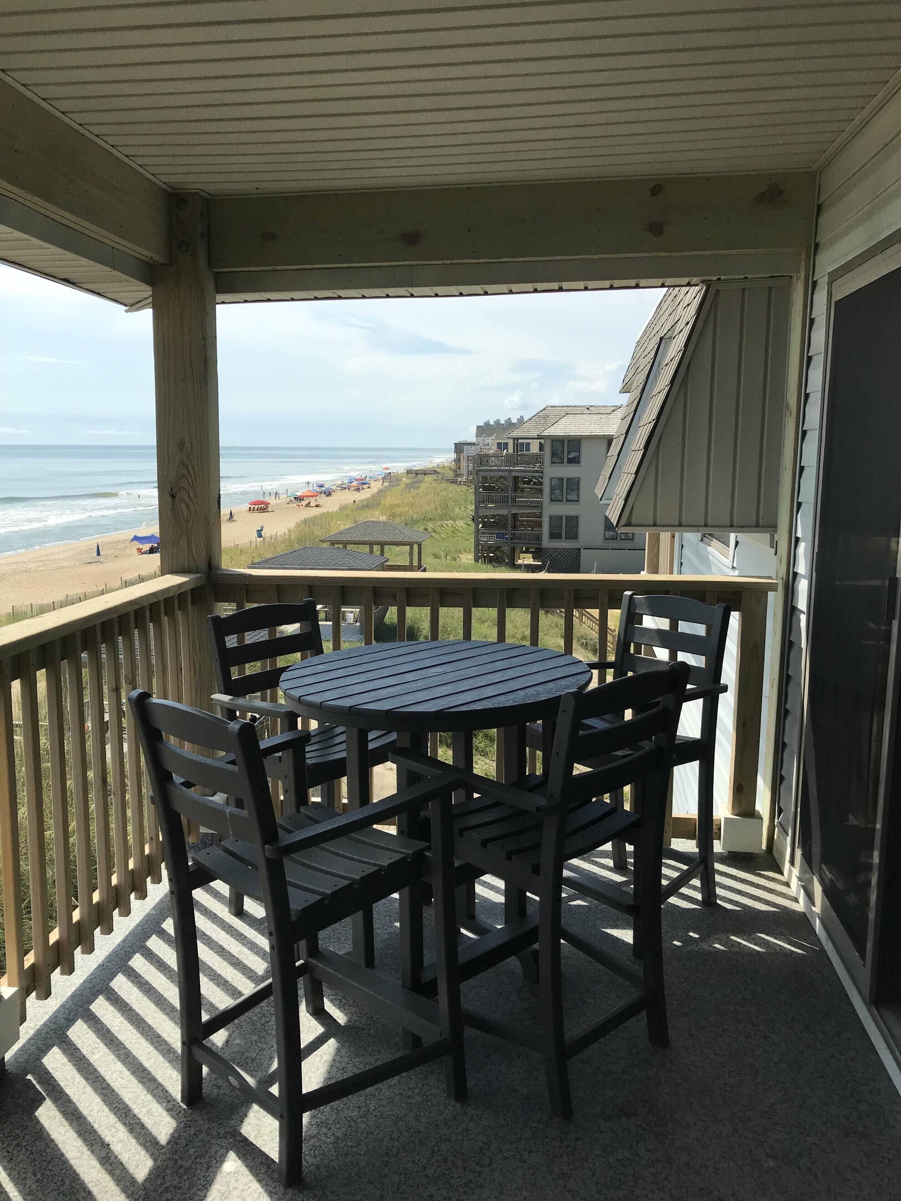 Un espace extérieur abrité, avec une table et des chaises, offrant une vue sur une plage et des bâtiments.