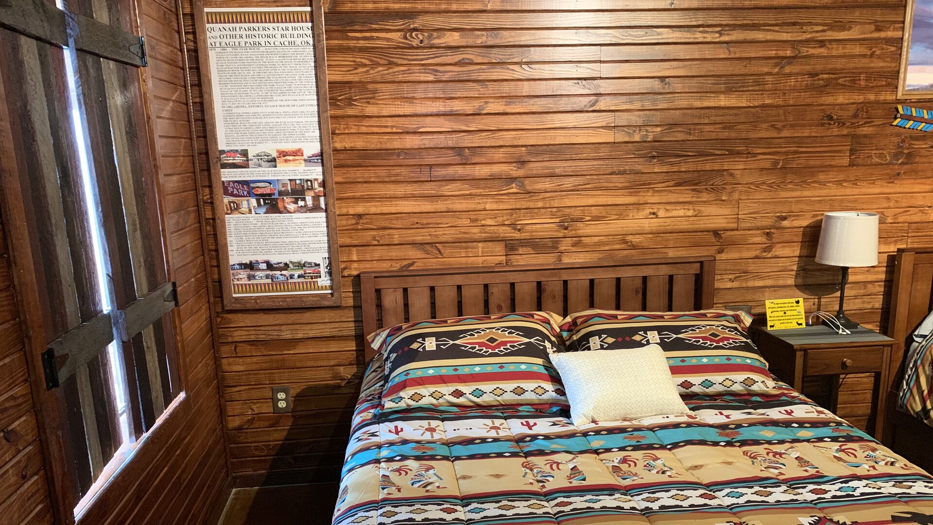 A wooden-paneled room with a bed, a nightstand, a lamp, and a poster on the wall.
