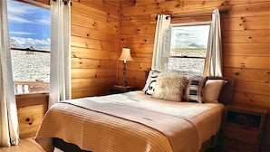 A wooden cabin bedroom with a bed, pillows, a nightstand, a lamp, and a window with curtains. - Enjoy a private mountain retreat at Paul's Cabin! (Hartsel)