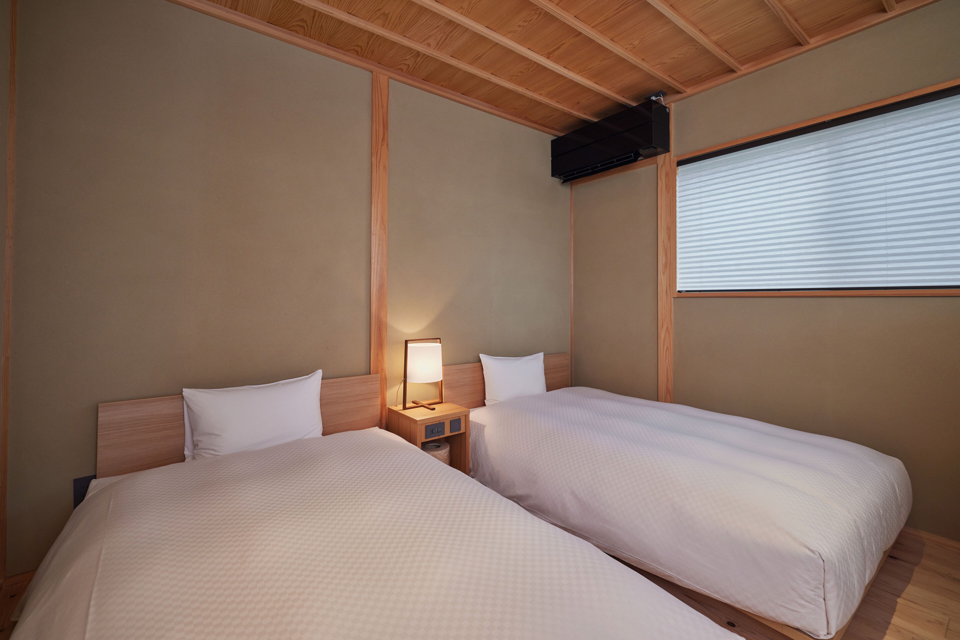 A hotel room with two beds, wooden headboards, a nightstand with a lamp, and a window with blinds.