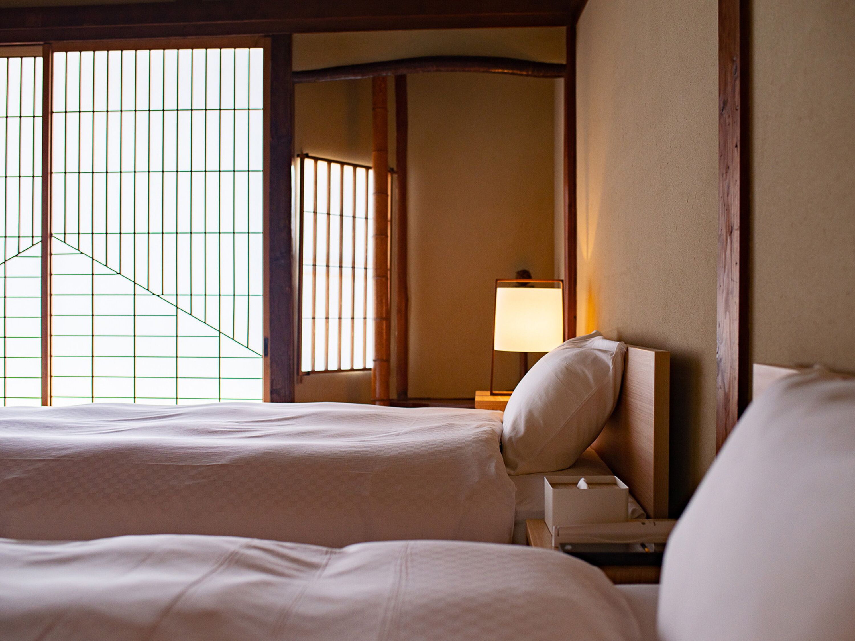 A hotel room with two beds, a nightstand with a lamp, and a view through a window with vertical blinds.
