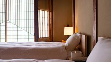 A hotel room with two beds, a nightstand with a lamp, and a view through a window with vertical blinds.