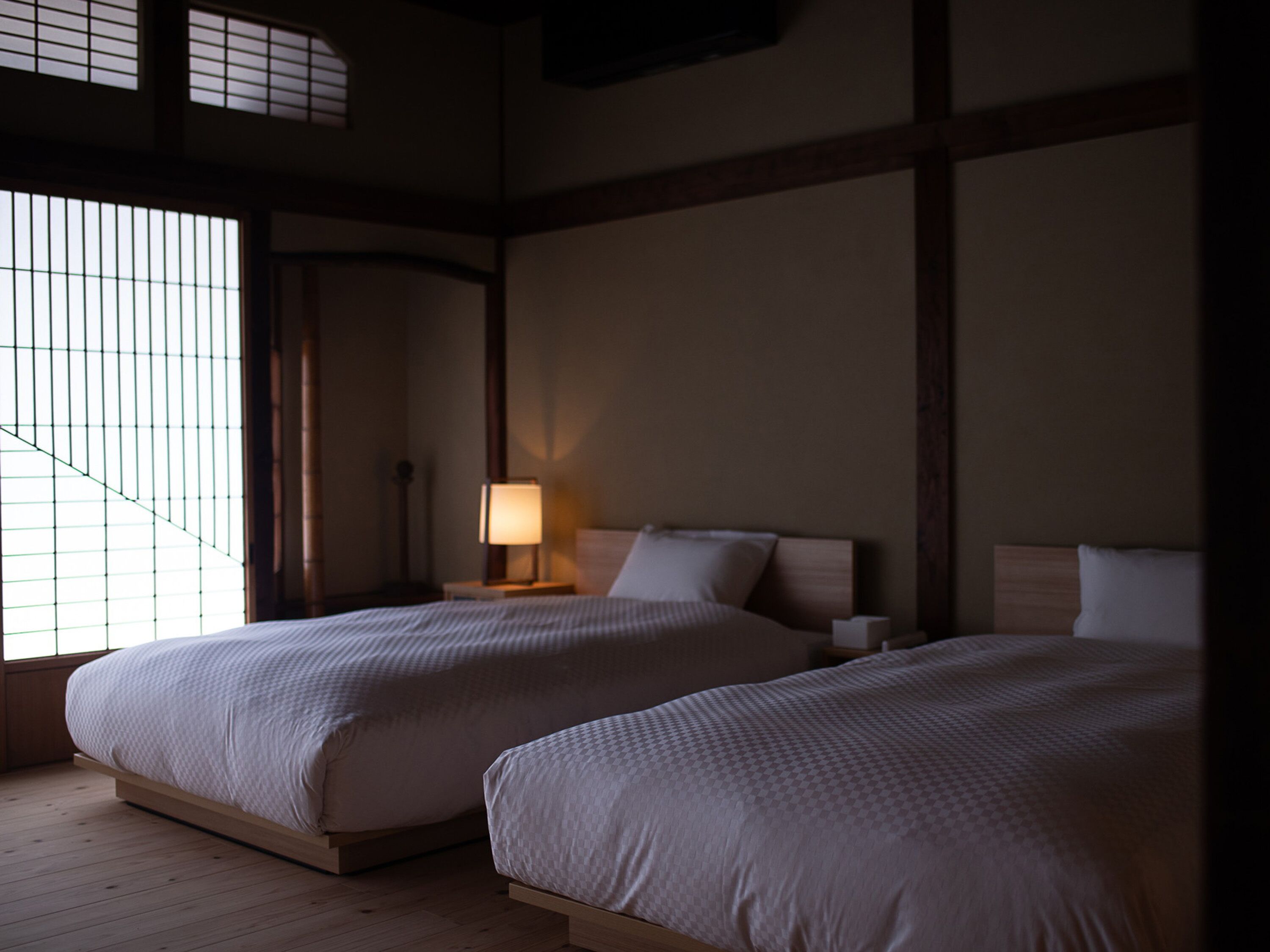 A room with two beds, a nightstand with a lamp, and a window with a view of the outside.