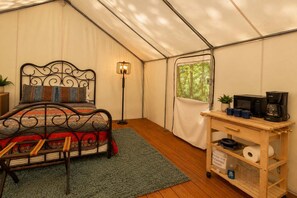 Une pièce aménagée comme une tente, avec un lit, une lampe sur pied, une fenêtre donnant sur les arbres et une petite table sur laquelle se trouvent une machine à café et des tasses.
