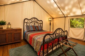 A tented room with a bed, a side table, a lamp, and a view of greenery outside.