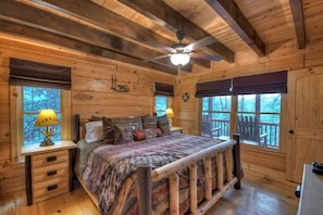 A wooden cabin bedroom with a bed, two bedside lamps, a ceiling fan, and large windows.
