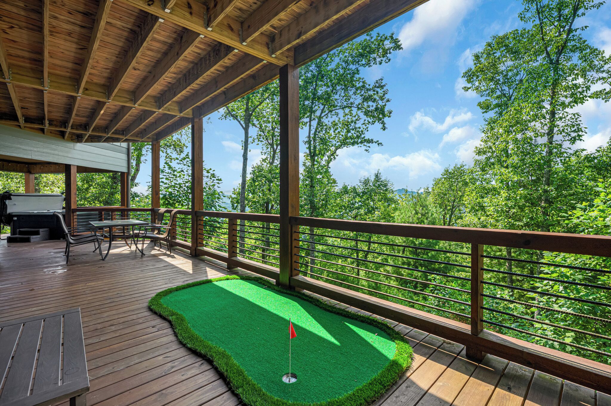 Una terraza de madera con un área de práctica de golf de césped artificial verde, mobiliario exterior y vistas a árboles.