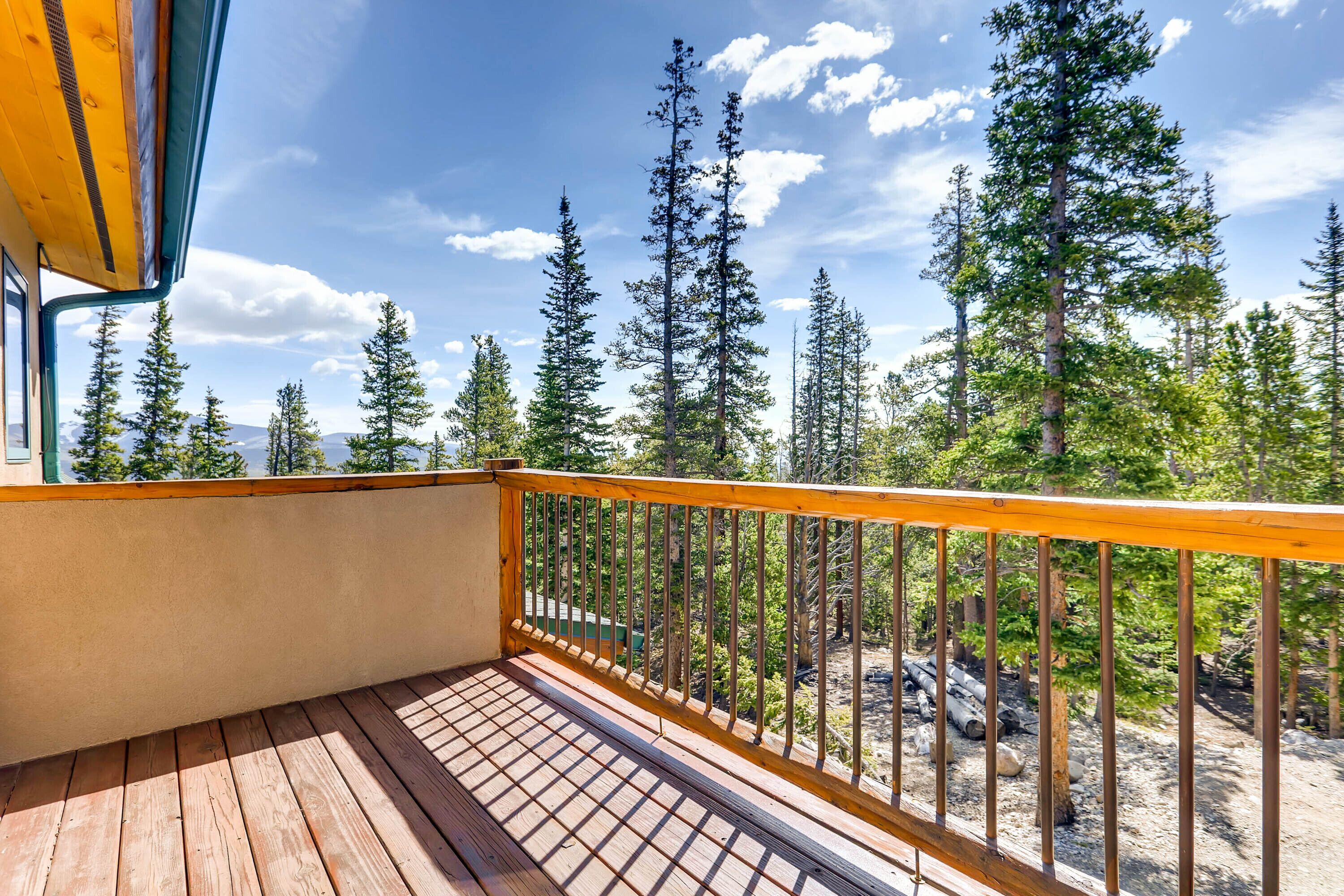 A wooden deck with a metal railing overlooking a forested area with tall trees and a clear sky.