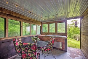 An enclosed patio area with a brick fireplace, a round table, and floral-patterned chairs. - Historic Cabin in Little Switzerland w/Fire Pit-Walk to village and shops! (Little Switzerland)
