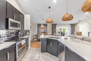 Een moderne keuken met een ingebouwde magnetron, fornuis en vaatwasser, een eethoek met tafel en stoelen, en een groot kookeiland met spoelbak.