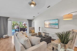 A modern living room with a sofa, armchairs, a coffee table, and a dining table. There is a large window with a view of the outdoors.