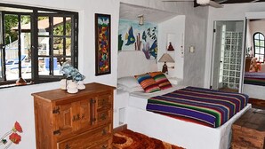 Un dormitorio con cama, cómoda de madera, ventana con persianas, un cuadro y una puerta que da a otra habitación.