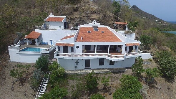 Una casa grande y blanca de dos pisos, con techo de tejas rojas, piscina y vista al mar.