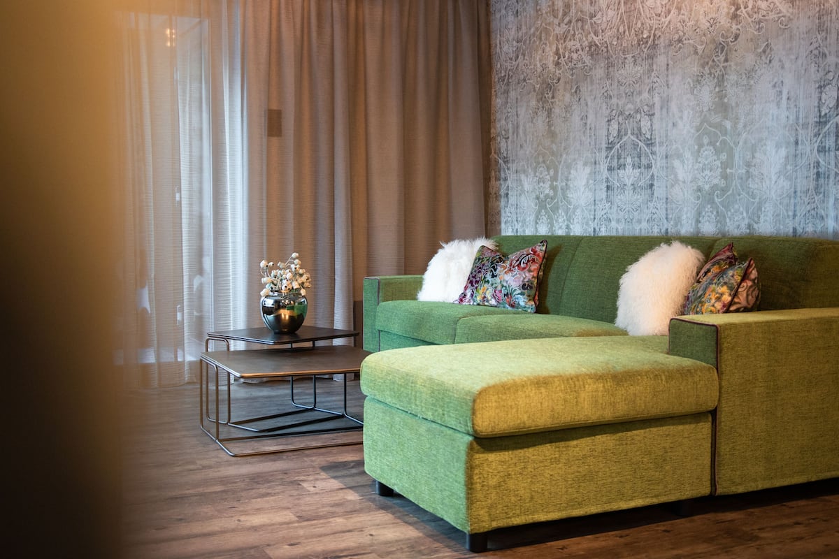 A green sofa with colorful pillows, a small side table with a vase, and a patterned wall.