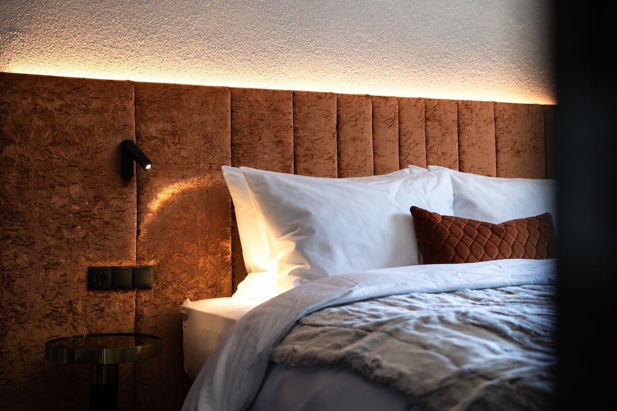 A bedroom with a wooden headboard, a bedside table with a lamp, and a bed with white bedding and a brown pillow.