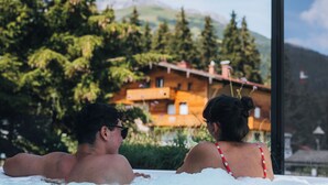 A couple relaxing in a hot tub with a mountain view and a wooden building in the background. - Apart Sailers (Sankt Anton am Arlberg)
