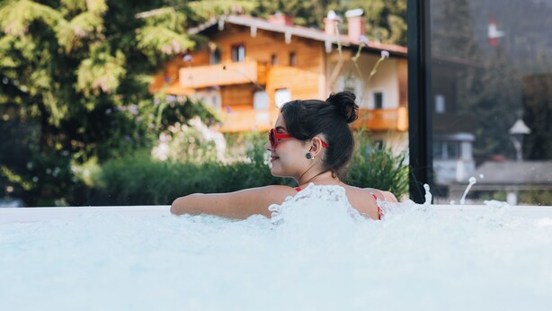 A person relaxing in a hot tub with a view of a wooden building and trees in the background. - Apart Sailers (Sankt Anton am Arlberg)