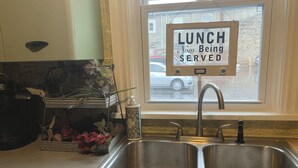 Une cuisine avec un évier en acier inoxydable, une fenêtre sur laquelle est affiché un panneau indiquant « DÉJEUNER – Service en cours » et une plante décorative posée sur le comptoir.