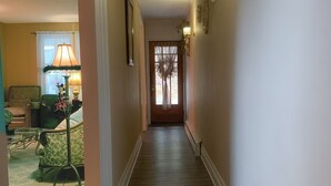 Un couloir doté d’un plancher en bois, d’un fauteuil recouvert d’un tissu vert, d’une lampe et d’une porte avec une fenêtre à l’extrémité.