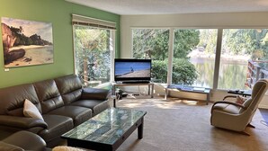 A living room with a brown leather sofa, a glass coffee table, a television, and a view of a lake through large windows.