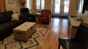 Un salon comprenant un canapé, des fauteuils, une table basse et un téléviseur. Il y a des fenêtres avec des stores, une porte et un tapis sur le sol.