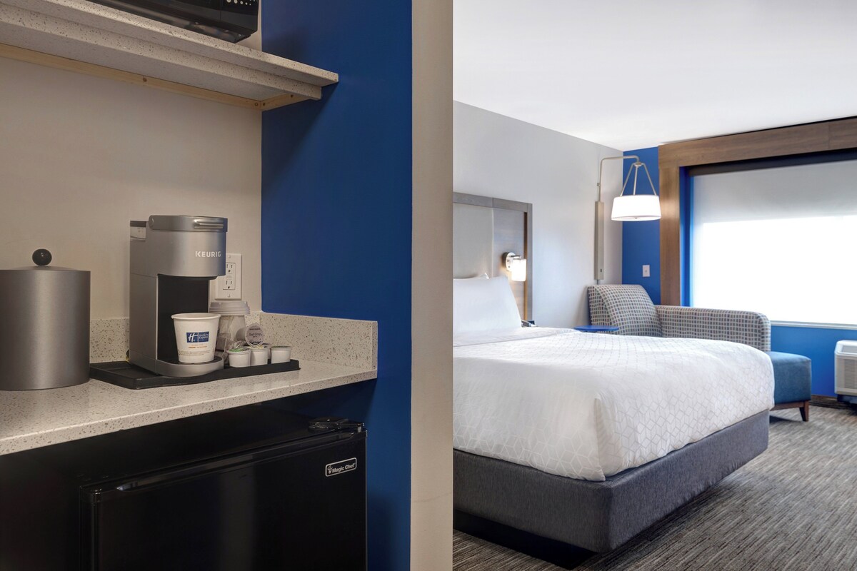 Habitación de hotel con kitchenette, una cama, mesita de noche, lámpara y ventana con vistas.