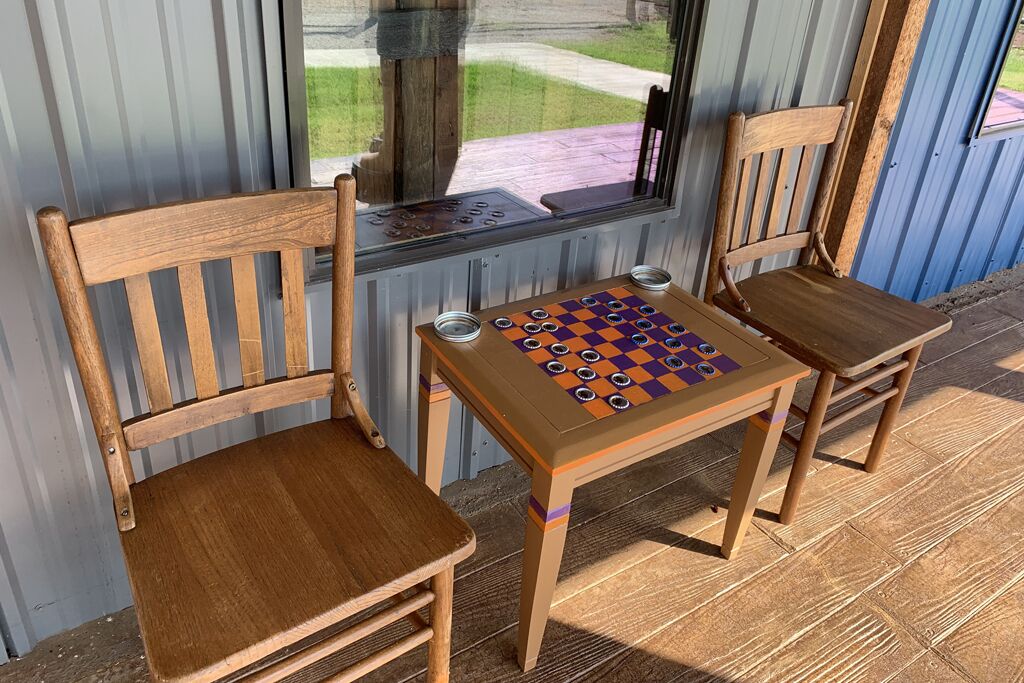 Una mesa de madera con un juego de mesa de cuadros, dos sillas de madera y una ventana que da a un jardín y un sendero.