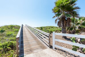 Un muelle de madera con barandillas blancas que conduce a una playa con palmeras y plantas propias del desierto.