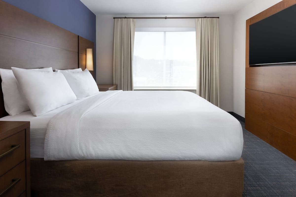 Une chambre d’hôtel avec un grand lit, une tête de lit en bois, une table de chevet, une télévision et une fenêtre avec des rideaux.