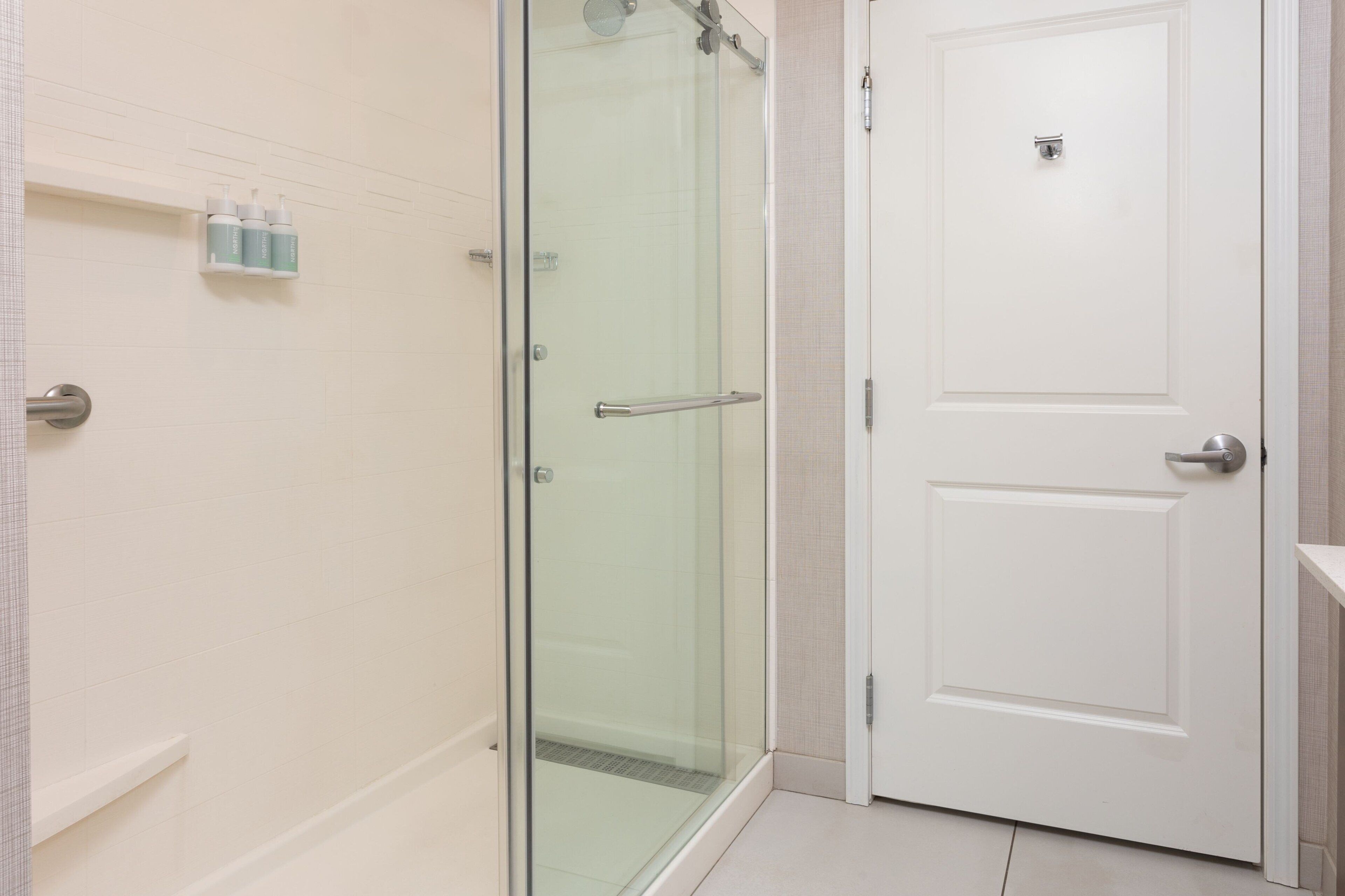 A modern bathroom with a glass shower enclosure, a white door, and a shelf with toiletries.