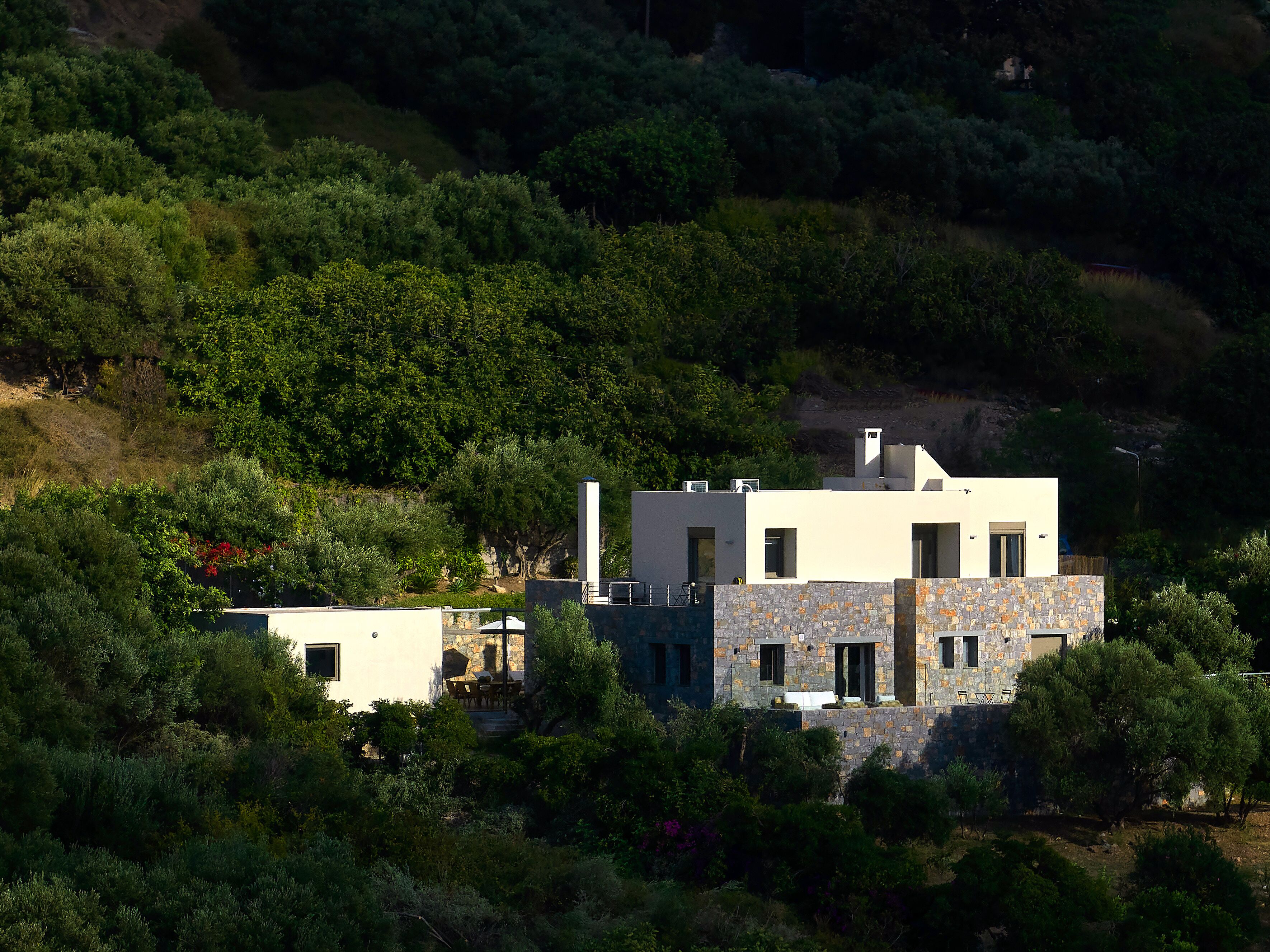 A modern two-story house with a stone facade, surrounded by dense greenery and hills.