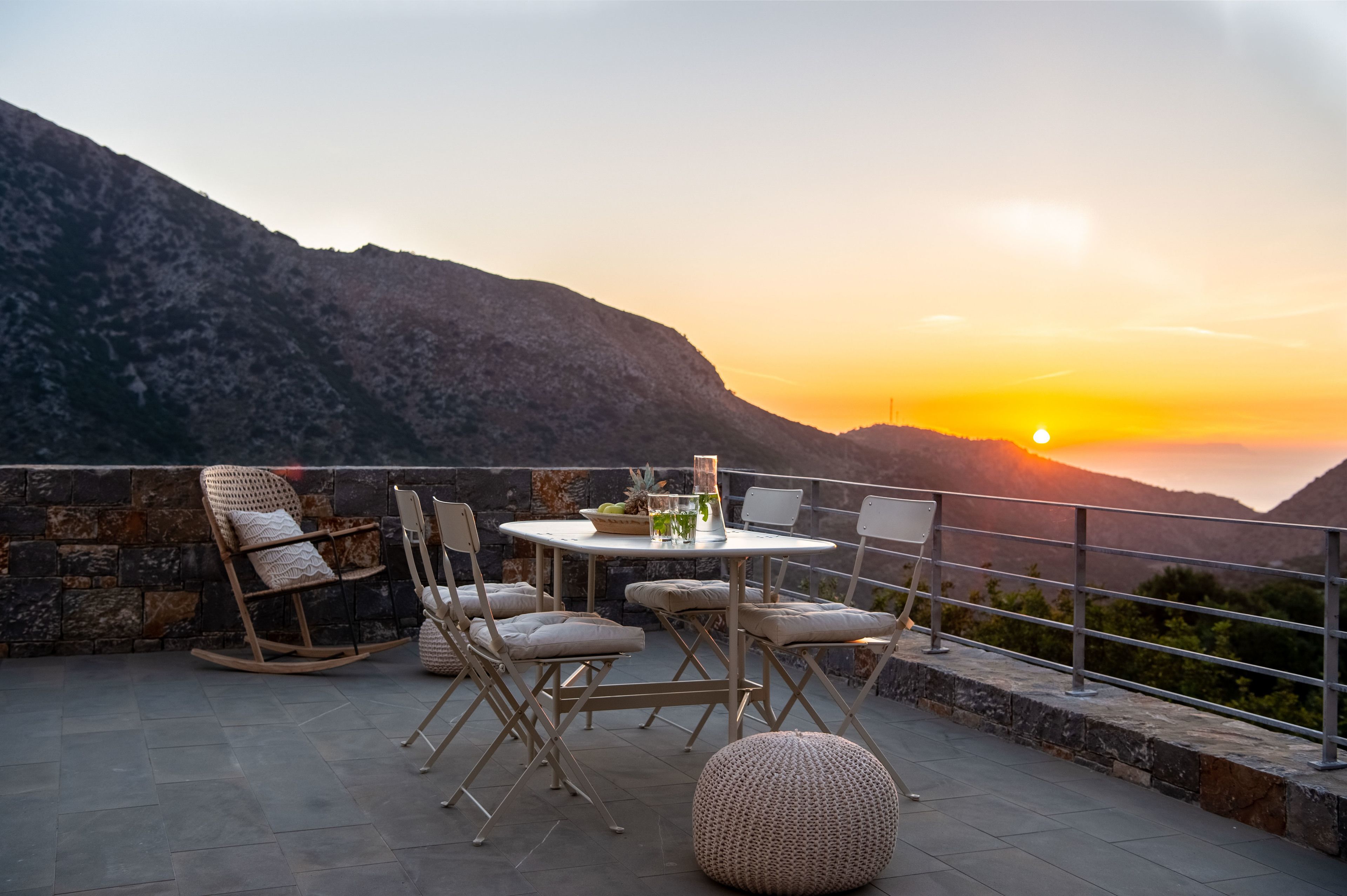 Uma varanda com mesa redonda, cadeiras e uma cadeira de balanço, com vista para uma paisagem montanhosa ao pôr do sol.