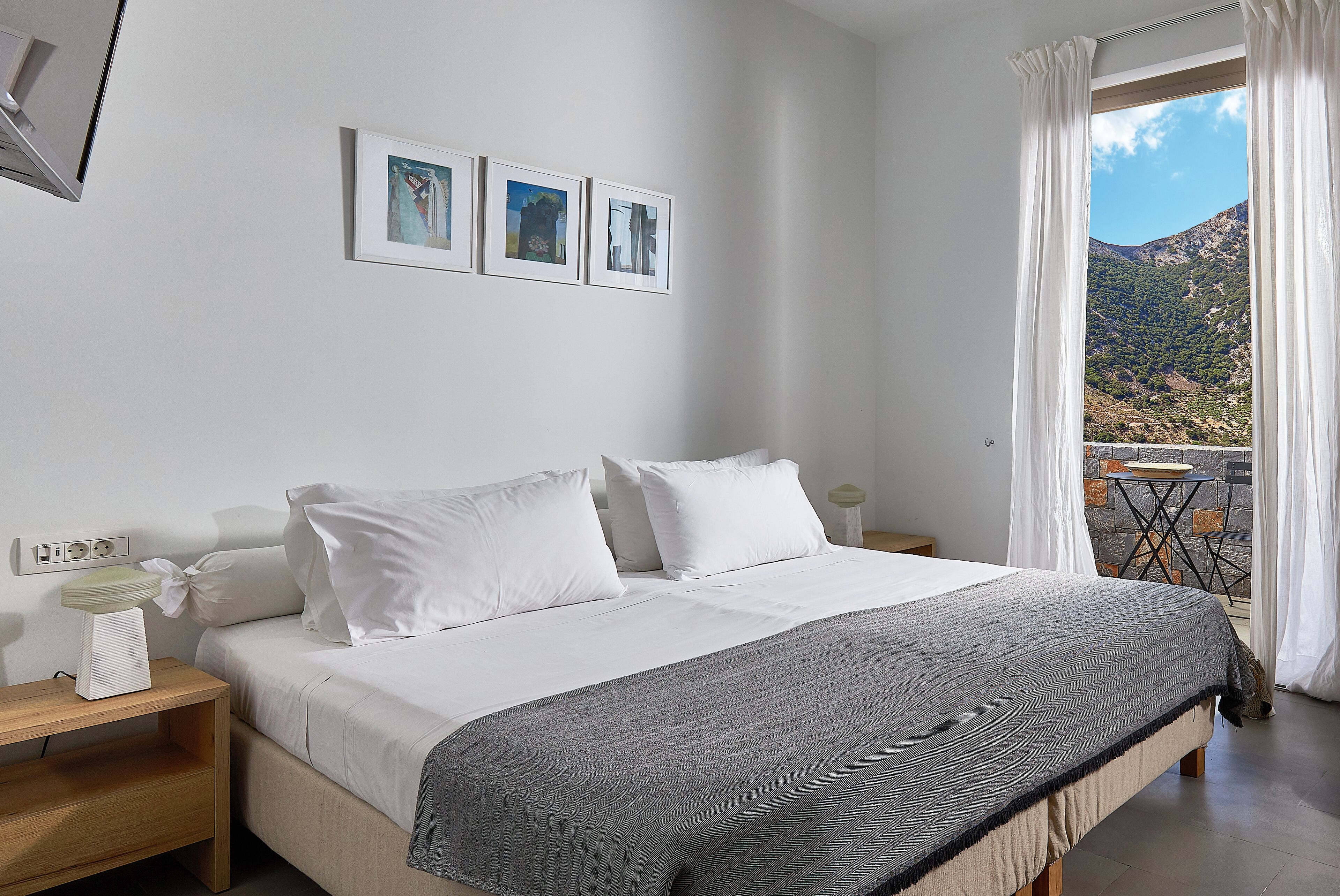 A bedroom with a bed, white pillows, a grey blanket, a nightstand, a bedside table, and a window with curtains.