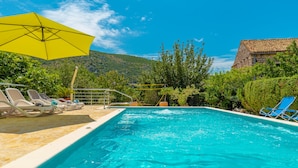 Piscina con sillas de descanso y una sombrilla amarilla, rodeada de vegetación y una montaña al fondo.