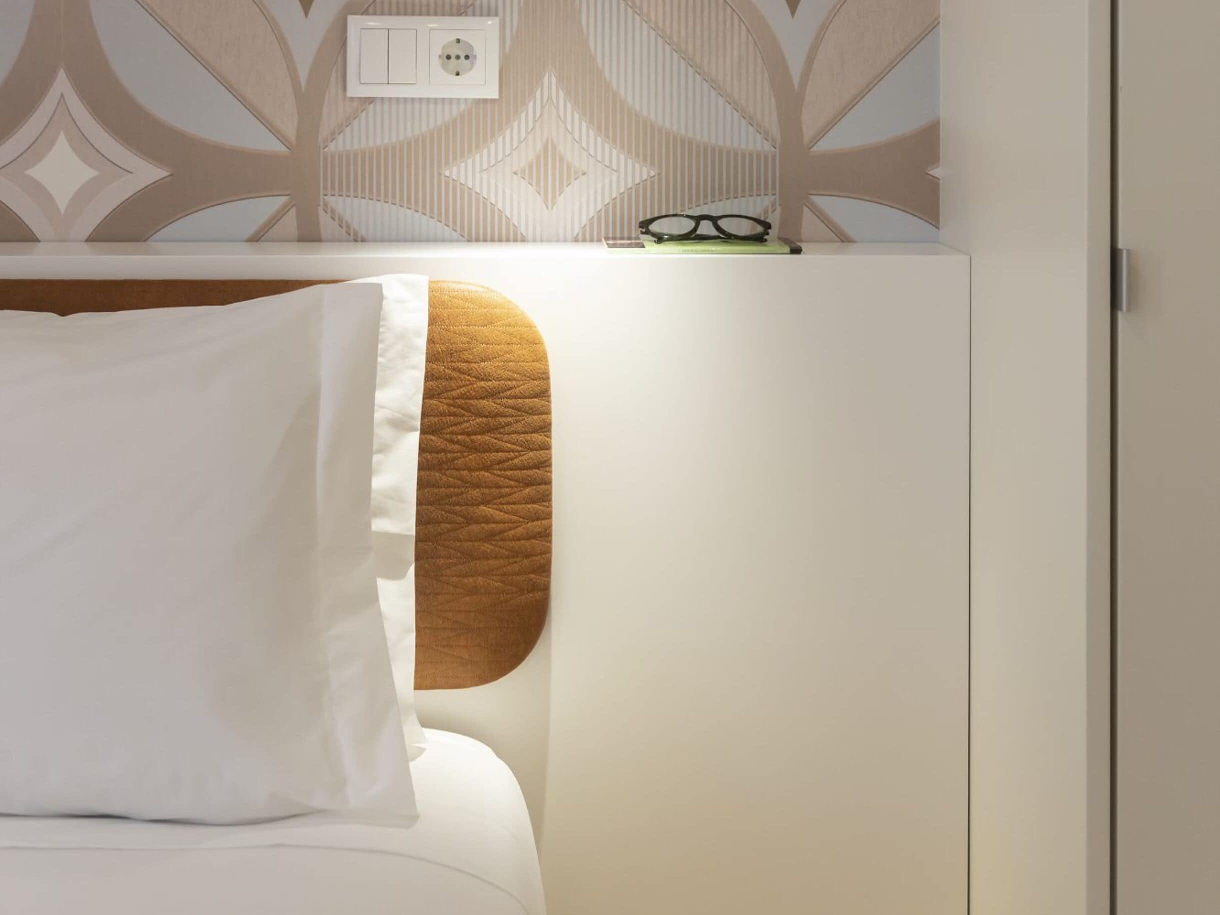 Un lit avec un oreiller blanc et une tête de lit marron, une prise de courant au mur et une paire de lunettes posées sur une étagère au-dessus du lit.