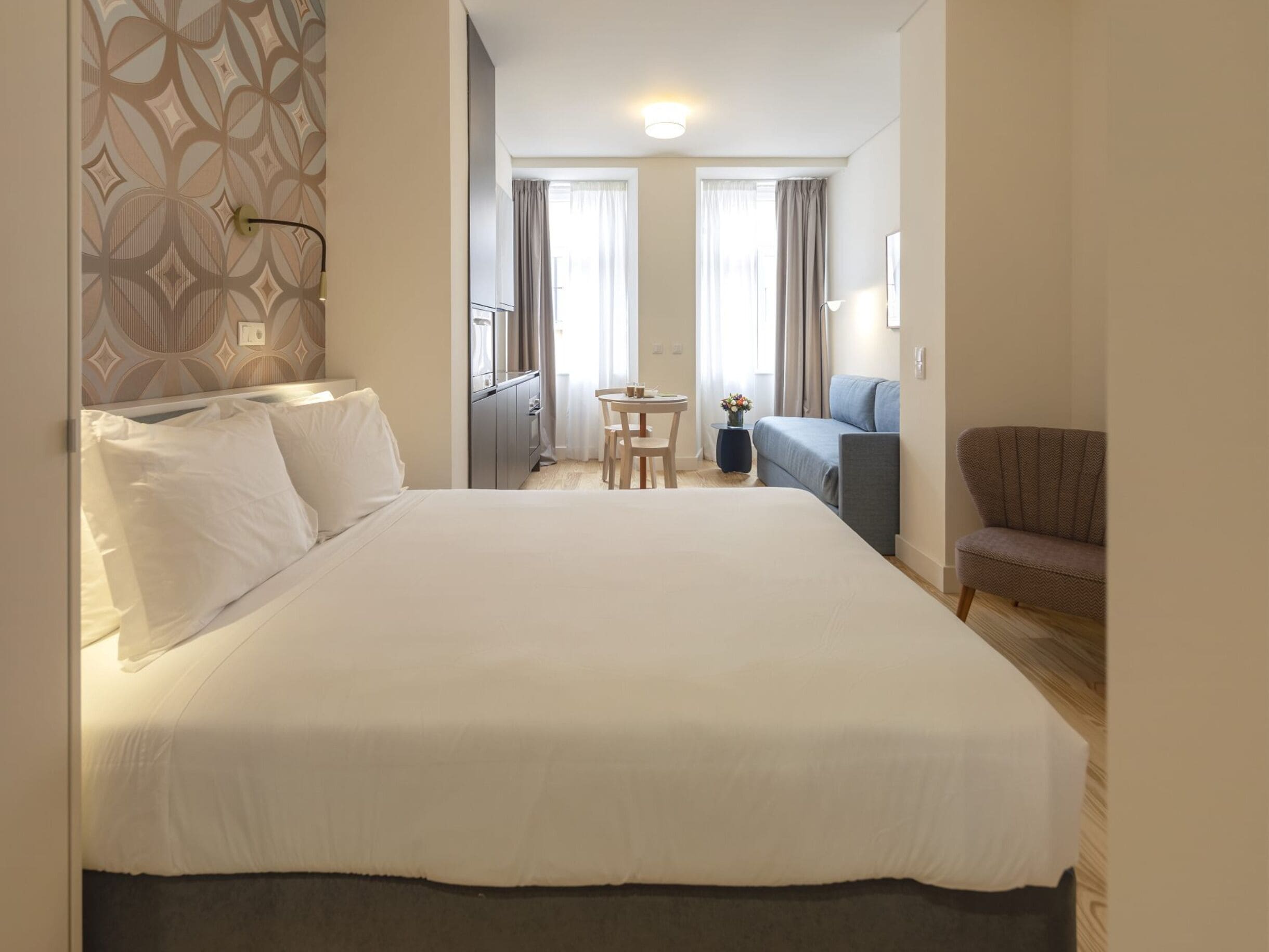 A hotel room with a large bed, a small table, a chair, a sofa, and a window with curtains.
