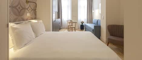 A hotel room with a large bed, a small table, a chair, a sofa, and a window with curtains.