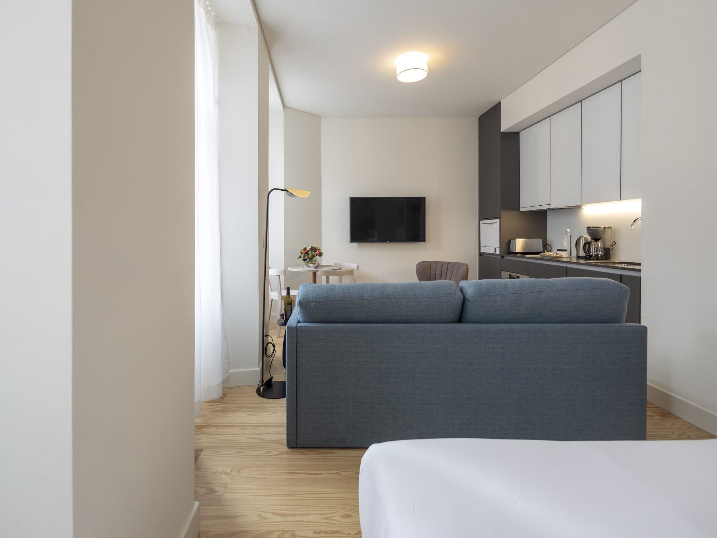 A modern hotel room with a grey sofa, a flat-screen TV, a dining area with a table and chairs, and a kitchenette with cabinets and a coffee machine.