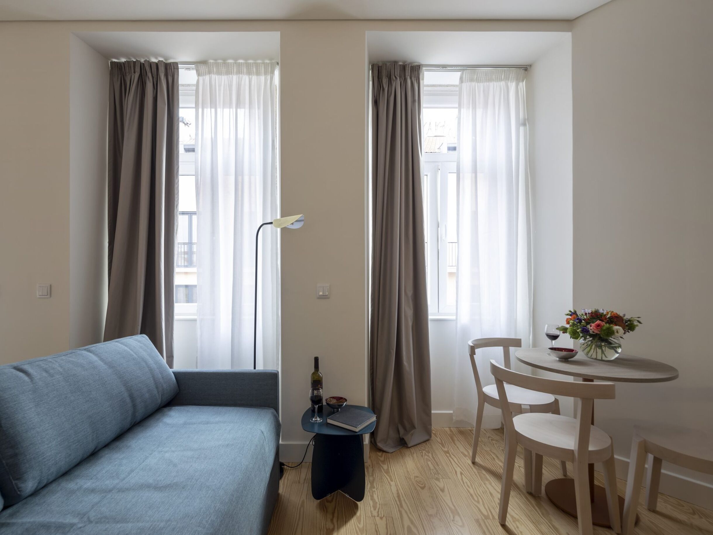 A modern living room with a sofa, a small table with chairs, a lamp, and a window with curtains.