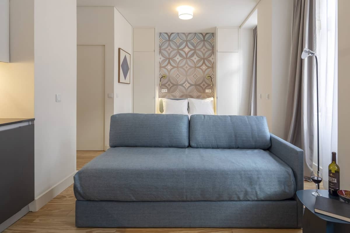 A modern bedroom with a grey sofa, a bedside table with a bottle and glass, and a patterned wallpaper.