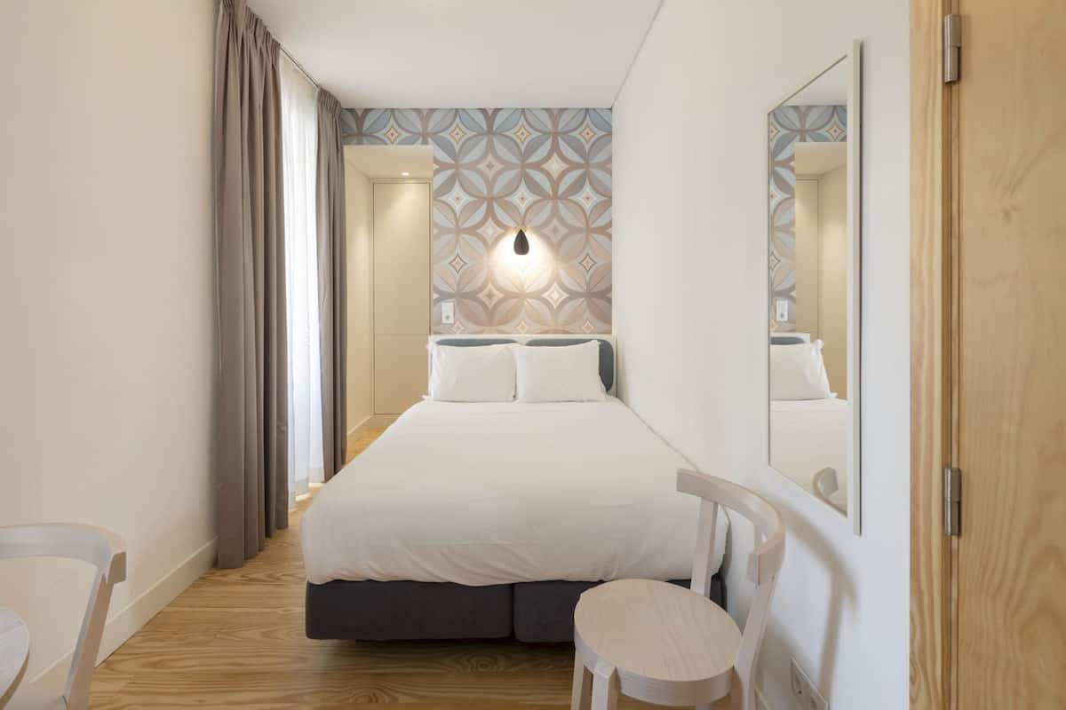 A hotel room with a large bed, a desk, a chair, a mirror, and patterned wallpaper.