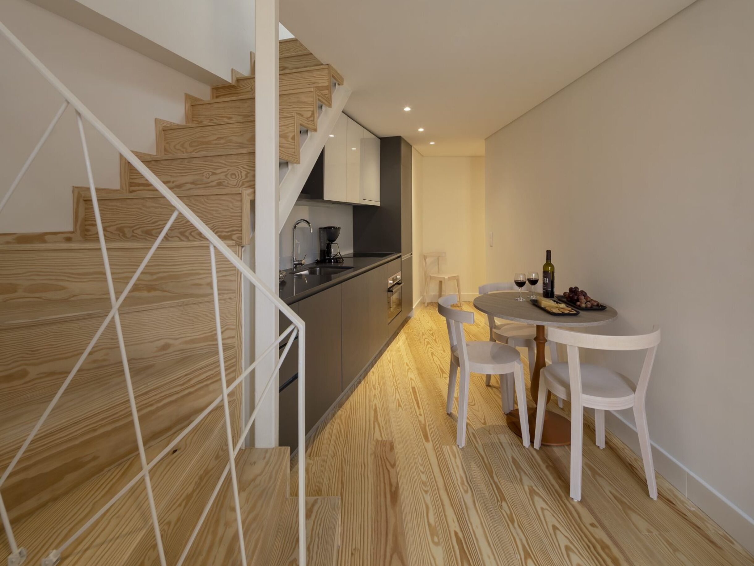 A modern kitchen with wooden flooring, a dining area with white chairs, and a staircase with wooden steps.