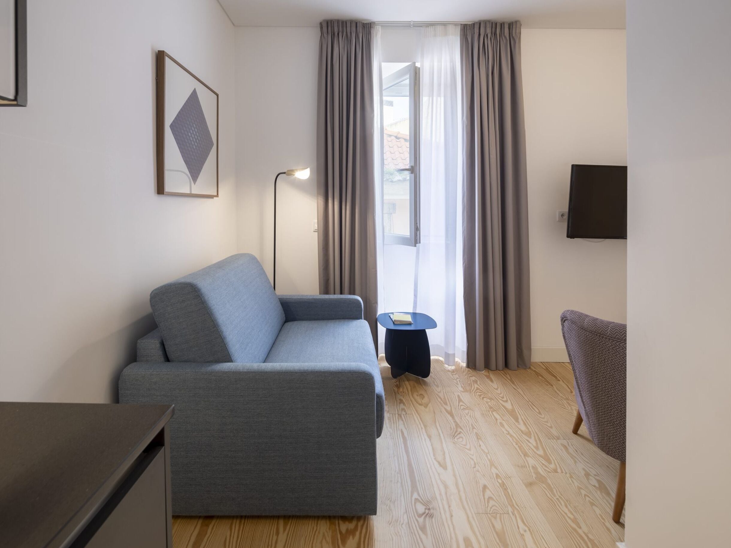 A modern living room with a grey sofa, a small side table, a chair, a floor lamp, and a TV mounted on the wall.