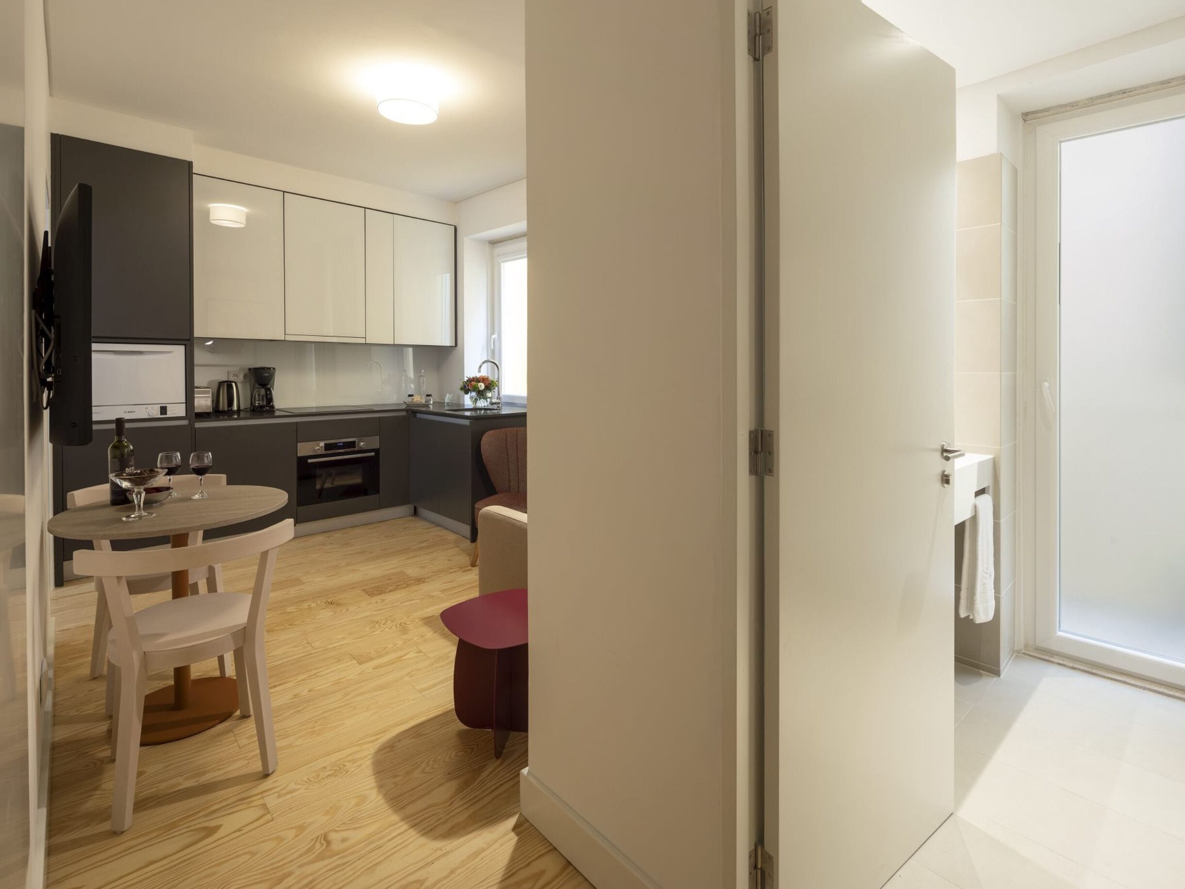 A modern kitchen with a dining area, wooden flooring, and a view through an open door.