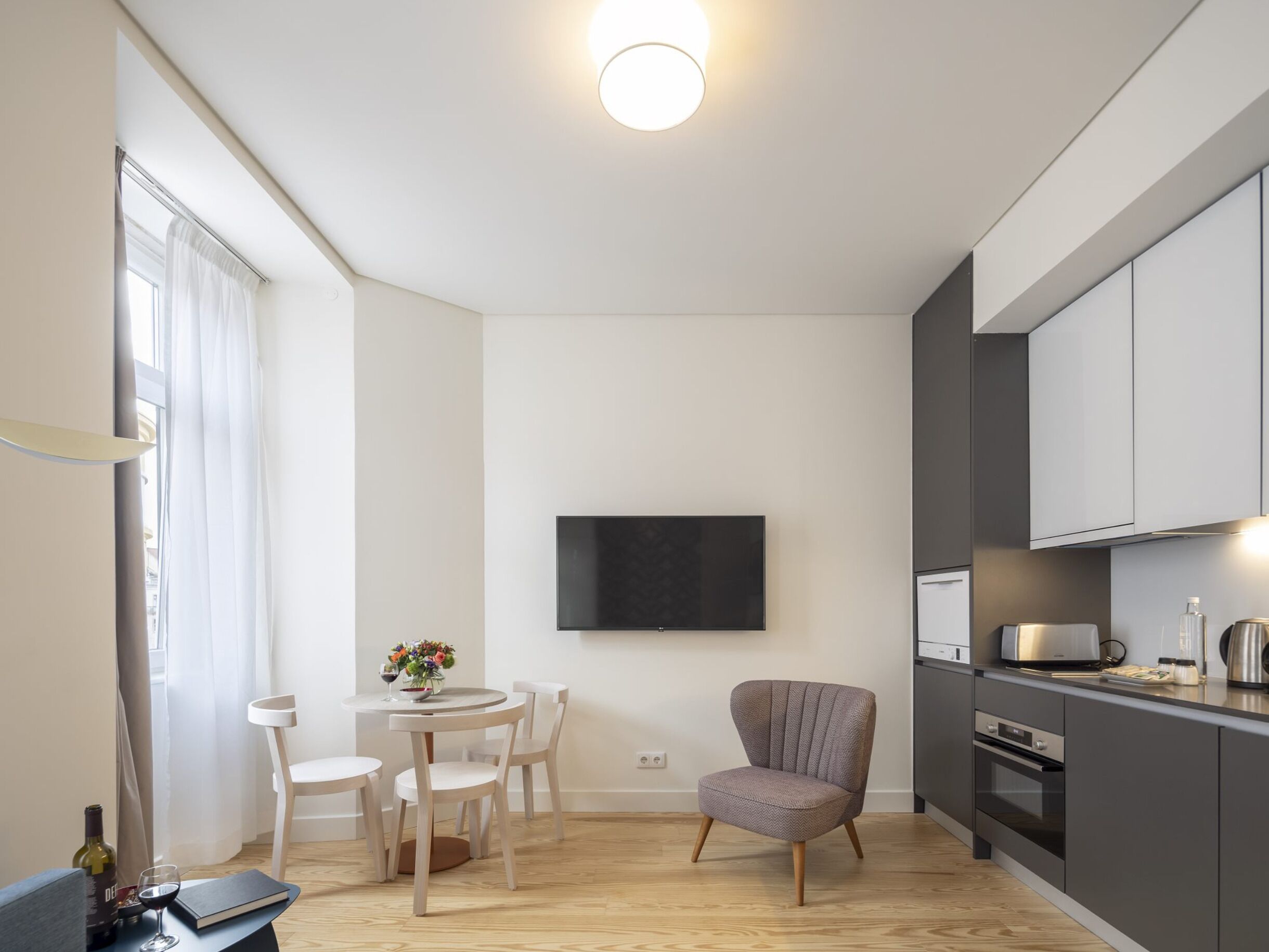 Une cuisine moderne équipée d’un téléviseur à écran plat, d’un coin repas avec une table et des chaises, et d’un fauteuil gris.