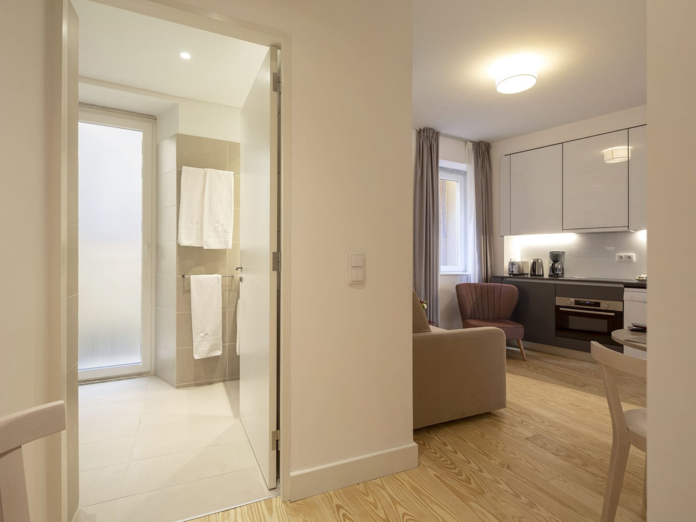 A modern hotel room with a wooden floor, a sofa, a dining table, a kitchen area, and a bathroom with a glass shower.