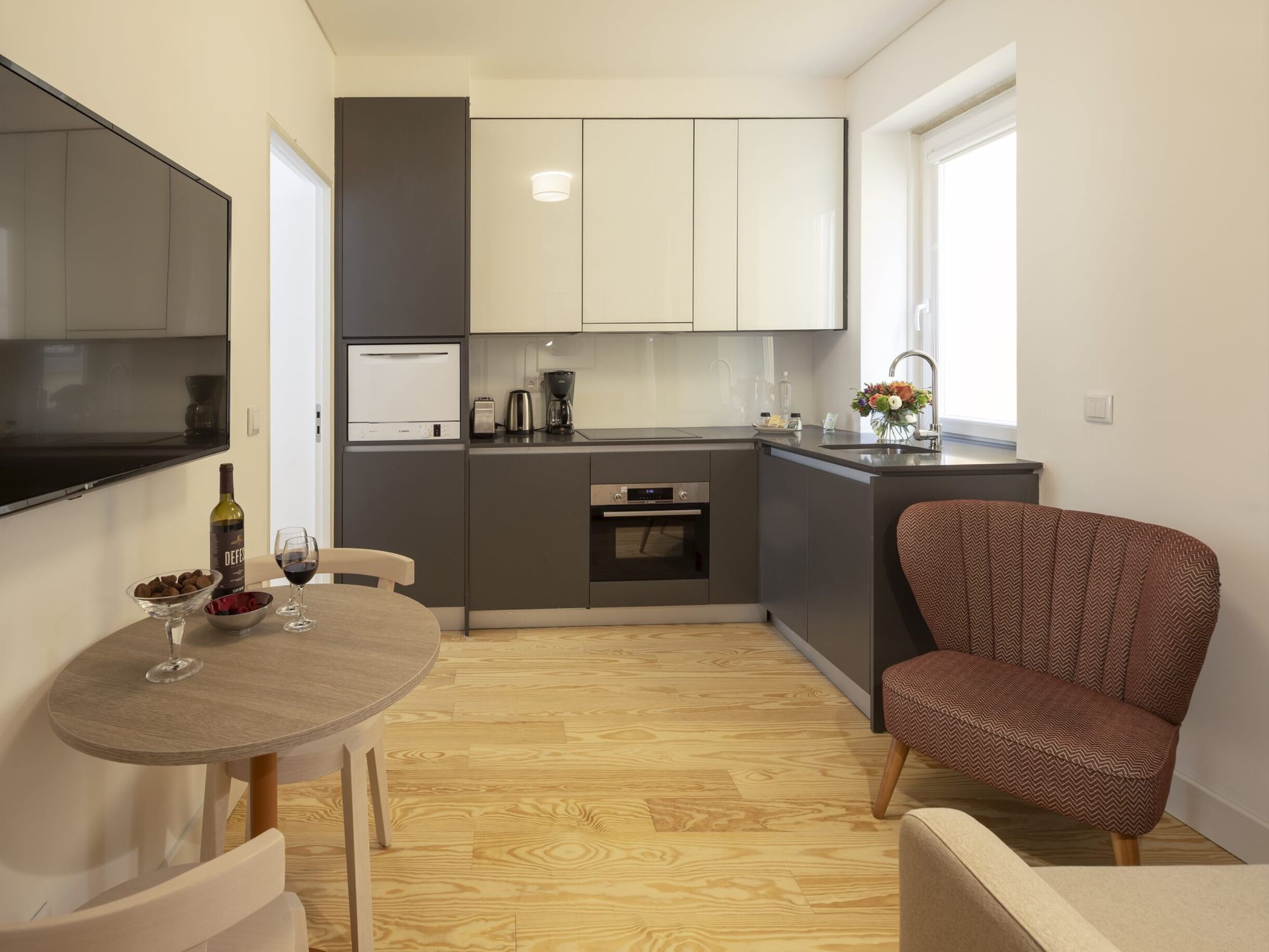 A compact kitchen with a dining area, a flat-screen TV, a wine bottle, and a chair.
