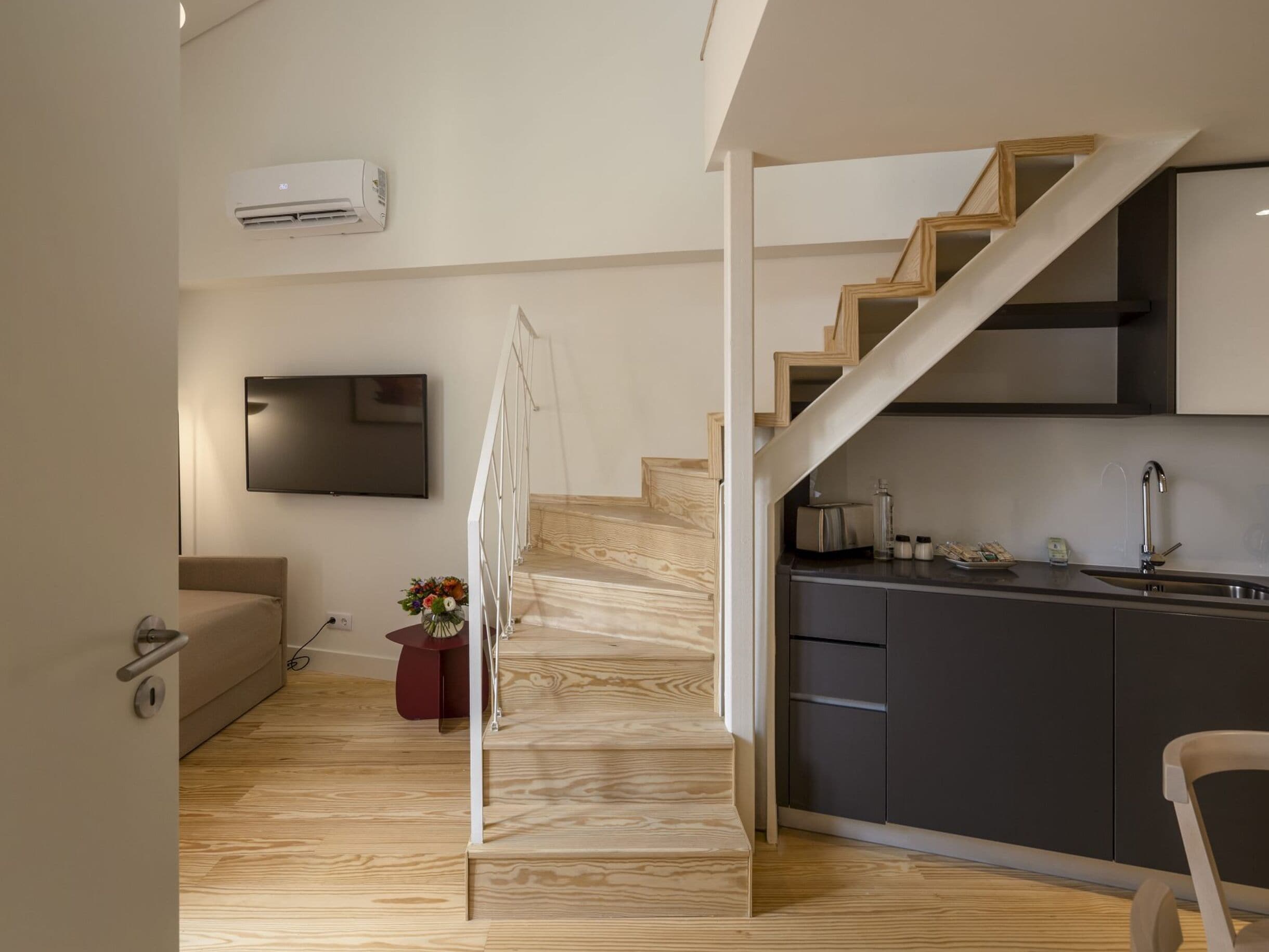 A modern kitchen with dark cabinetry, a wooden staircase, a flat-screen TV mounted on the wall, and a small dining area with a chair.