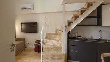 A modern kitchen with dark cabinetry, a wooden staircase, a flat-screen TV mounted on the wall, and a small dining area with a chair.