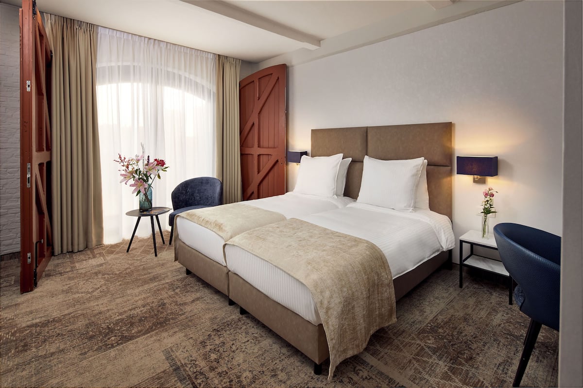 Ein Hotelzimmer mit einem großen Bett, einem kleinen Tisch, einem Sessel, einem Nachttisch mit Lampe und einem großen Fenster mit Vorhängen.